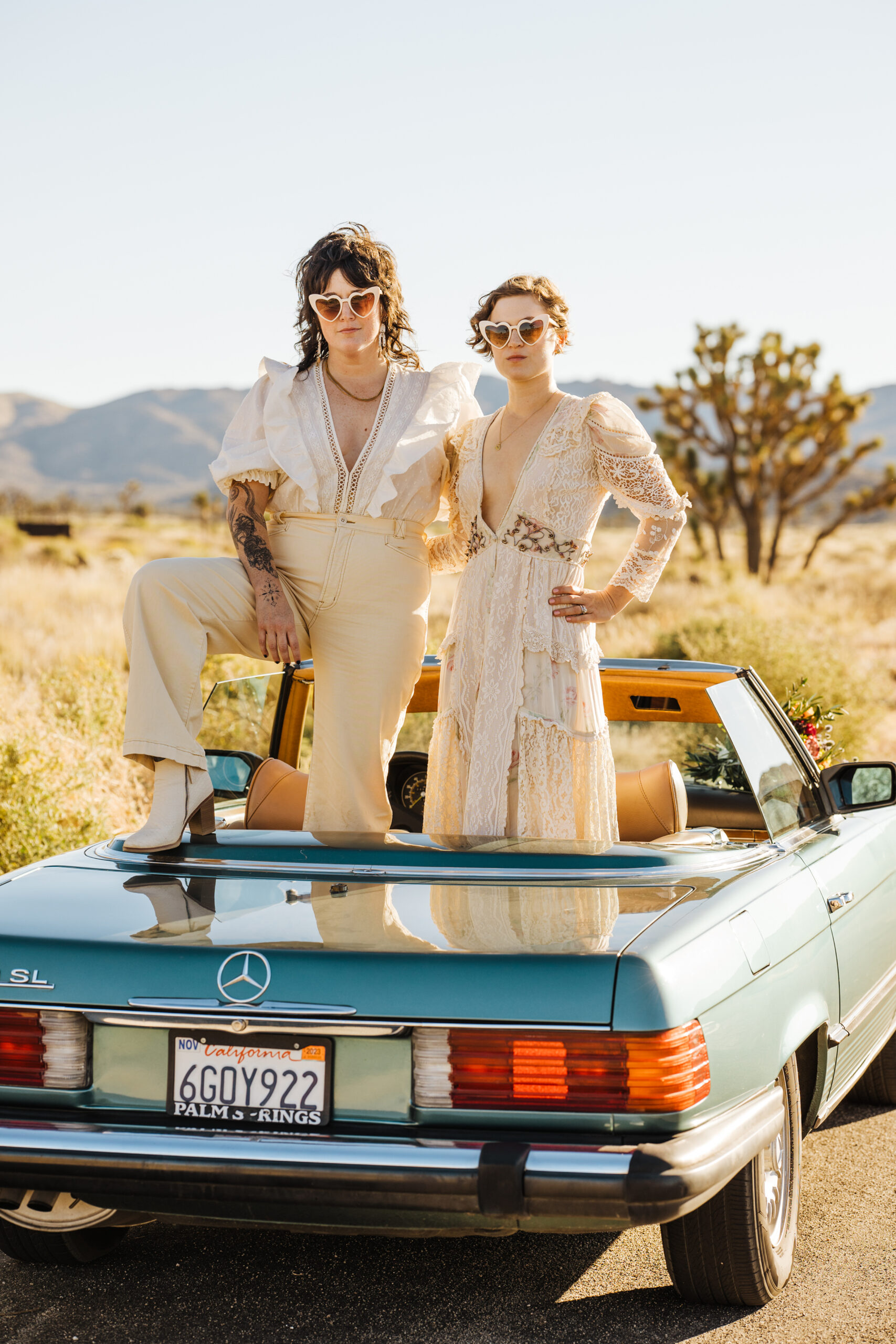 Two brides eloping in Joshua Tree standing in a vintage convertible car with sunglasses